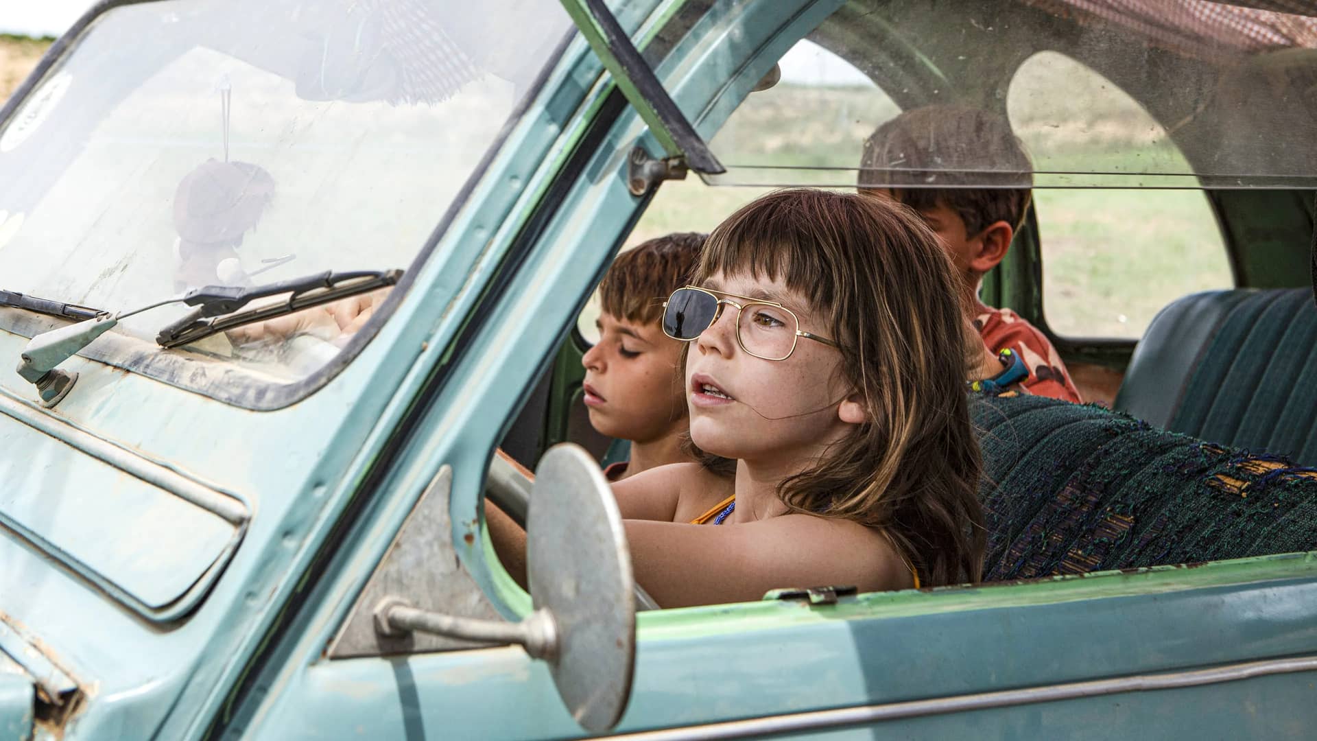 Una niña con unas gafas de sol rotas conduciendo un coche abandonado que es parte de la pelicula de Alcarras